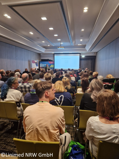 Eine Gruppe Menschen sitzen in einem Vortrag oder Workshop vor einer Leinwand. Vorne steht eine Referentin.